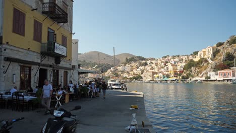 Gente-En-El-Restaurante-Y-Motocicletas-Pasando-Por-La-Calle-Angosta,-Casas-Pintorescas-En-La-Colina-Al-Fondo,-Puerto-De-La-Isla-Symi.