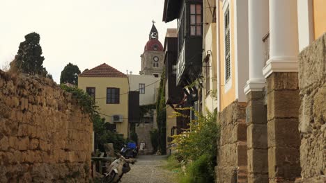 Narrow-street-with-stone-walls-and-traditional-houses-in-Old-Town-of-Rhodes,-Clock-tower-in-the-background