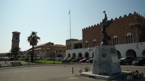 Estatua-De-La-Victoria,-El-Almirante-Periklis-Ioannidis-Junto-Al-Palacio-Del-Gobernador,-La-Torre-De-La-Iglesia-Evangelista-Al-Fondo