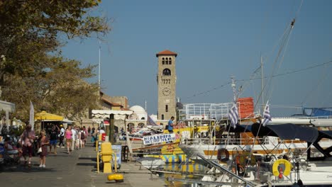 Colorful-harbour-of-Rhodes-Town,-Tourboats,-Tourists-and-the-clock-tower-of-the-Evangelist-Church-in-background