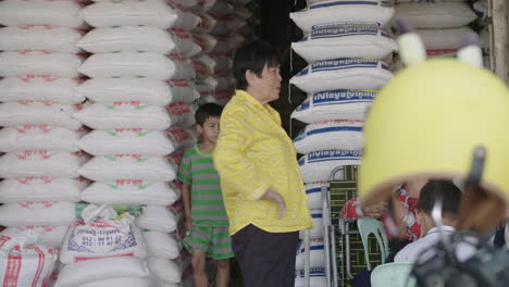 Niños-Y-Mujeres-Mayores-Frente-A-Una-Gran-Cantidad-De-Bolsas-De-Arroz-Grandes-En-Un-Puesto-En-El-Mercado-De-Arroz-Al-Aire-Libre-En-Camboya