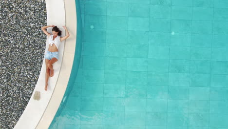 Vista-Aérea-De-Arriba-Hacia-Abajo-De-Una-Mujer-Joven-Posando-Junto-A-Una-Piscina