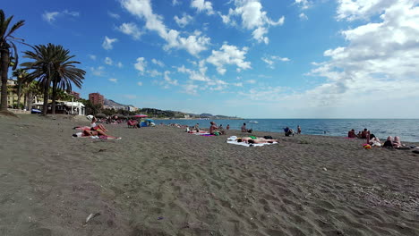 Middle-Age-People-relaxing-on-sandy-beach-with-palm-tree-near-Mediterranean-sea,-blue-water-during-the-day