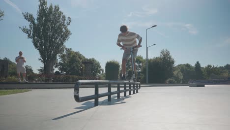 Hombre-En-Scooter-Acrobático-Realiza-Deslizamiento-En-Pasamanos-Metálicos-De-Skatepark,-Día-Soleado-De-Verano