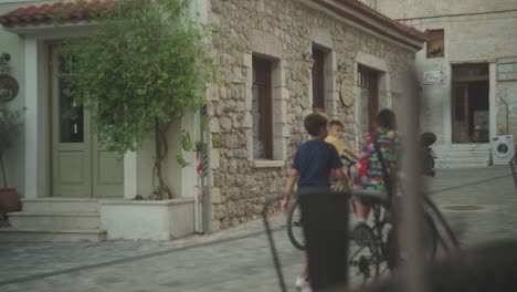 Eine-Reisende-Frau-Geht-Die-Straße-Hinauf,-Während-Kinder-Vor-Einem-Alten-Backsteingebäude-In-Einer-Mediterranen-Stadt-Fahrrad-Fahren