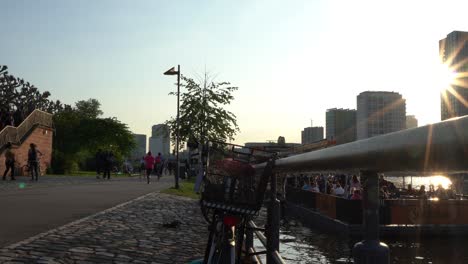 Puesta-De-Sol-En-Frankfurt-Con-Gente-Caminando-Junto-Al-Río,-Silueta-De-Una-Bicicleta-En-Primer-Plano,-Paisaje-Urbano-Al-Fondo