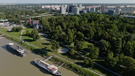 Vista-Aérea-Por-Drones-Del-Triste-Janka-Krala---Parque-A-Orillas-Del-Río-Danubio-Con-árboles-Verdes-Y-Casas-De-Apartamentos-En-La-Parte-Trasera-En-Un-Día-Soleado