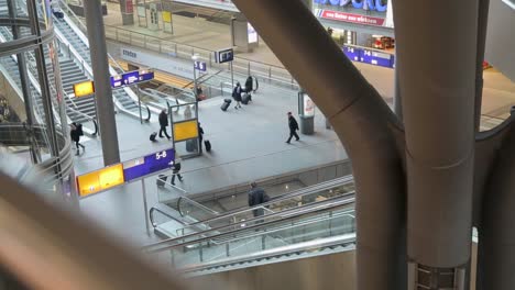 Elevated-view-of-a-bustling-train-station-with-passengers-walking-and-escalators-in-motion,-indoor-daylight-scene