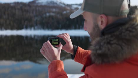 Young-bearded-man-in-orange-jacket-holding-an-action-camera-and-taking-photo-of-winter-landscape