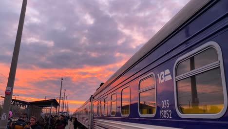 Zug-Der-Ukrainischen-Eisenbahn-Ukrzaliznycia-Mit-Wunderschönem-Rosafarbenen-Sonnenaufgangshimmel-Am-Bahnhof-Chelm-In-Polen,-Ukrainische-Flüchtlinge-Warten-Am-Frühen-Morgen-Darauf,-In-Den-Zug-Nach-Kiew-In-Der-Ukraine-Einzusteigen,-4k-Kippt-Nach-Unten