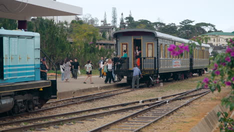 Da-Lat-Railway-Station---Old-Locomotive-Train-Travels-on-Tracks-while-People-Enter-Ancient-Train-Wagon,-Groups-of-Tourists-walk-on-Platform