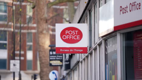 Un-Autobús-Rojo-Pasa-Al-Fondo-De-Un-Cartel-De-La-Oficina-De-Correos,-Una-Instalación-Pública-Que-Proporciona-Correo-Y-Otros-Servicios