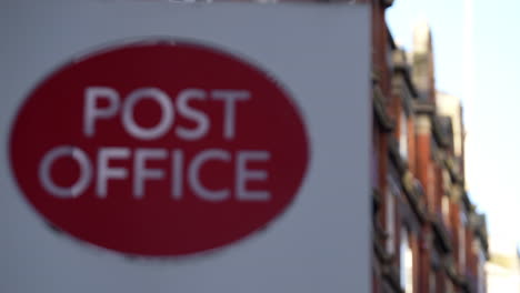 A-red-brick-ornate-building-stands-behind-a-red-and-white-Post-Office-logo-on-a-sign,-a-public-facility-that-provides-mail-and-other-services