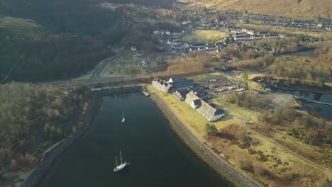 Toma-Aérea-De-Arriba-Hacia-Abajo-Del-Hotel-Isles-Of-Glencoe-Con-Un-Velero-En-El-Lago-Leven-En-Las-Tierras-Altas-De-Escocia