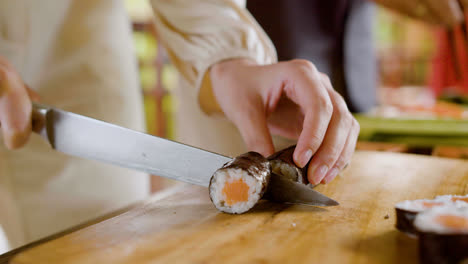 Close-up-view-of-hands-of-a-woman-making-sushi-on-top-of-a-kitchen-board.-On-the-background-her-partner-is-cutting-other-ingredients