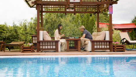 Distant-view-of-asian-couple-eating-ramen-while-they-are-talking-sitting-on-a-japanese-garden-porch-near-a-swimming-pool