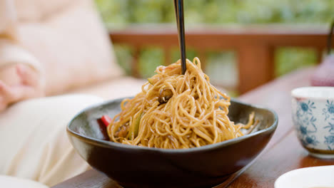 Close-up-view-of-hands-of-a-woman-picking-up-noodles-with-chopsticks-from-the-plate-on-a-table-outdoors