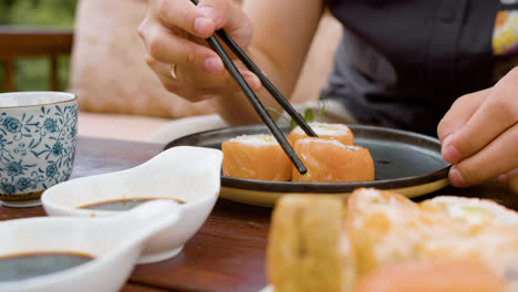 Close-up-view-of-hands-of-a-man-picking-up-pieces-of-sushi-with-chopsticks-and-dipping-them-in-soy-sauce-on-a-table-outdoors