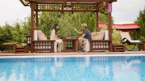 Distant-view-of-asian-couple-eating-ramen-while-they-are-talking-sitting-on-a-japanese-garden-porch-near-a-swimming-pool