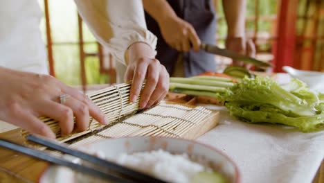 Vista-De-Cerca-De-Las-Manos-De-Una-Pareja-Asiática-Haciendo-Sushi-Y-Cortando-Aguacate-Sobre-Una-Tabla-De-Cocina-En-Casa