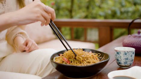 Close-up-view-of-hands-of-a-woman-picking-up-noodles-with-chopsticks-from-the-plate-on-a-table-outdoors