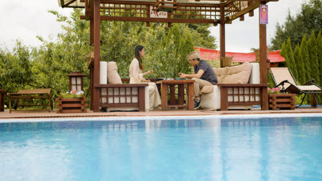 Distant-view-of-asian-couple-eating-ramen-while-they-are-talking-sitting-on-a-japanese-garden-porch-near-a-swimming-pool