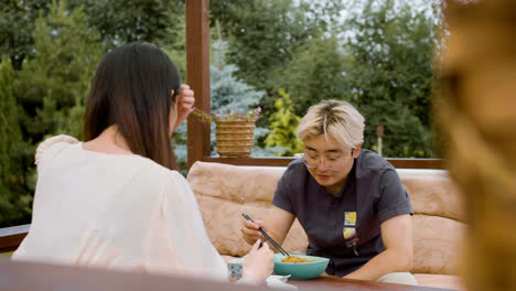 Vista-Trasera-De-Una-Mujer-Asiática-Comiendo-Ramen-Mientras-Habla-Con-Su-Pareja-Sentada-En-El-Porche-De-Un-Jardín-Japonés.