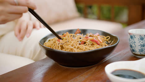 Close-up-view-of-hands-of-a-woman-picking-up-noodles-with-chopsticks-from-the-plate-on-a-table-outdoors