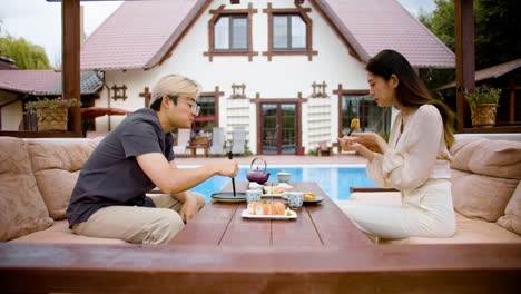 Side-view-of-asian-couple-eating-sushi-while-they-are-talking-sitting-on-a-japanese-garden-porch