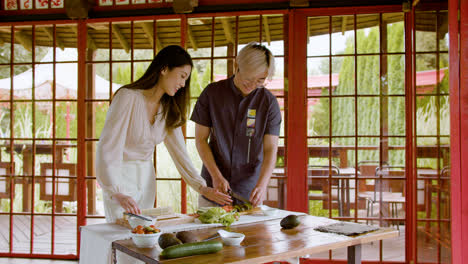 Vista-De-Cerca-De-Una-Pareja-Asiática-Haciendo-Sushi-Y-Cortando-Aguacate-Sobre-Una-Tabla-De-Cocina-Mientras-Hablan-En-Casa