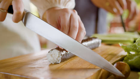 Vista-De-Cerca-De-Las-Manos-De-Una-Mujer-Haciendo-Sushi-Sobre-Una-Tabla-De-Cocina.-En-El-Fondo,-Su-Pareja-Está-Cortando-Aguacate.