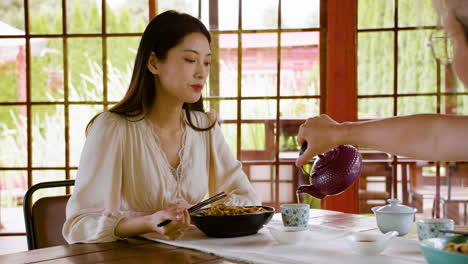 Asian-woman-eating-ramen-while-her-partner-is-pouring-tea-in-cups-while-they-are-sitting-at-a-table-in-a-japanese-home