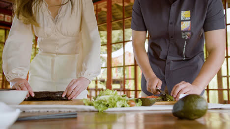 Vorderansicht-Eines-Asiatischen-Paares,-Das-Zu-Hause-Sushi-Zubereitet-Und-Avocado-Auf-Einem-Küchenbrett-Schneidet