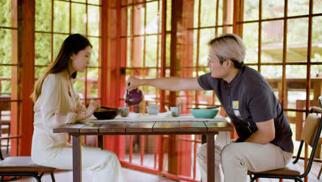 Side-view-of-a-woman-eating-ramen-while-her-partner-is-pouring-tea-in-cups-while-they-are-sitting-at-a-table-in-a-japanese-home