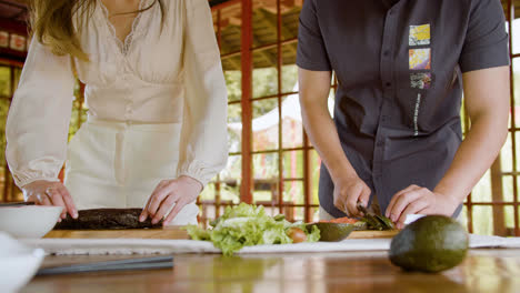 Front-view-of-asian-couple-hands-making-sushi-and-cutting-avocado-on-top-of-a-kitchen-board-at-home