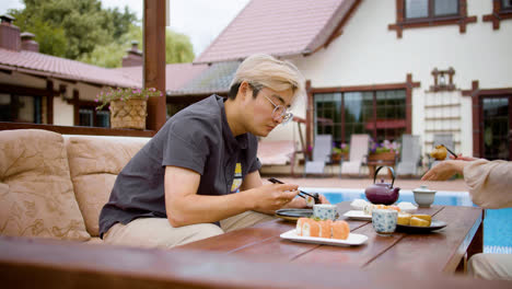 Side-view-of-asian-man-eating-sushi-while-she-is-talking-with-her-partner-sitting-on-a-japanese-garden-porch