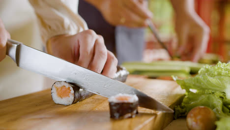 Close-up-view-of-hands-of-a-woman-making-sushi-on-top-of-a-kitchen-board.-On-the-background-her-partner-is-cutting-avocado