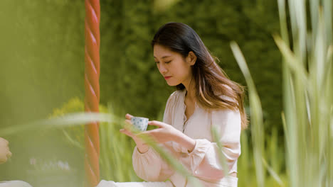 Vista-Lejana-De-Una-Mujer-Japonesa-Con-Cabello-Largo-Bebiendo-Té-De-Una-Taza-En-Un-Jardín-Japonés