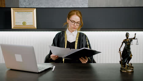 Kaukasische-Rechtsberaterin-In-Toga-Sitzt-An-Einem-Tisch-Im-Büro-Und-Liest-Ein-Dokument.-Auf-Dem-Tisch-Stehen-Ein-Computer-Und-Eine-Statue-Der-Gerechtigkeit.