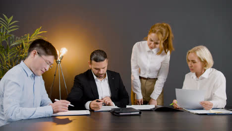 Group-of-multiethnic-legal-advisors-talking-and-reading-documents-sitting-at-a-table-in-the-office.-One-of-them-is-standing-between-two-co-workers