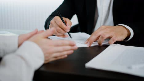 Close-up-view-of-the-hands-of-caucasian-man-writing-on-a-document-at-the-table-in-the-office.-His-costumer-is-making-gestures-to-explain-something-to-him