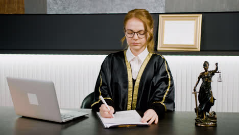 Caucasian-female-legal-advisor-wearing-toga-while-writing-on-a-document-sitting-a-table-in-the-office.-There-is-a-computer-and-a-statue-of-justice-on-the-table.