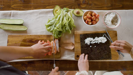 Vista-Superior-De-Las-Manos-De-Una-Mujer-Haciendo-Sushi-Sobre-Una-Tabla-De-Cocina.-En-El-Fondo,-Su-Pareja-Está-Cortando-Pescado-Fresco.