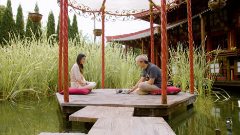 Japanese-couple-walking-over-the-bridge-of-a-Japanese-garden.-Then-they-stop-on-the-porch-and-sit-on-caushions