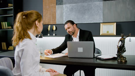 Caucasian-legal-advisor-sitting-at-a-table-with-laptop-in-the-office.-He-is-advising-a-ginger-woman