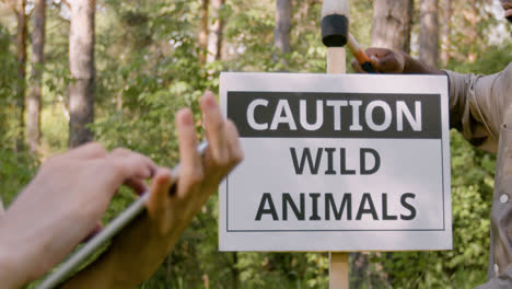 Vista-De-Cerca-Del-Guardabosques-Afroamericano-Instalando-Un-Letrero-Para-Advertir-Que-Hay-Animales-Salvajes-En-El-Bosque-Mientras-Su-Compañera-De-Trabajo-Usa-Una-Tableta