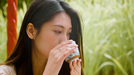 Close-up-view-of-japanese-woman-with-long-hair-drinking-tea-from-a-cup-while-sitting-on-a-japanese-garden-porch