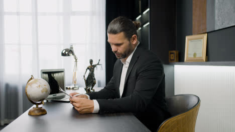 Side-view-of-caucasian-male-legal-advisor-wearing-a-black-suit-reading-a-document-and-sitting-a-table-in-the-office