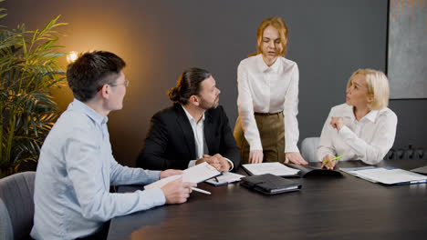 Group-of-multiethnic-legal-advisors-talking-and-reading-documents-sitting-at-a-table-in-the-office.-One-of-them-is-standing-between-two-co-workers