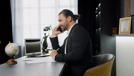 Side-view-of-caucasian-male-legal-advisor-wearing-a-black-suit-talking-on-the-phone-and-sitting-a-table-in-the-office